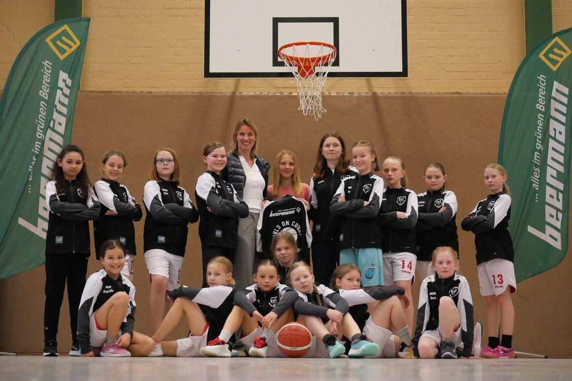 Gruppenfoto eines Basketballteams mit Trainerinnen, in Sportkleidung, vor einem Basketballkorb in einer Halle.