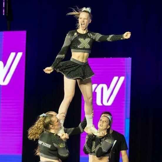 Cheerleaderin wird von zwei Teamkolleginnen in die Luft gehalten, w&auml;hrend eine weitere Person im Hintergrund zuschaut.