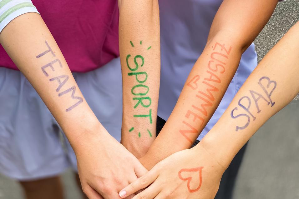 Vier Hände mit bunten Schriftzügen "TEAM", "SPORT", "ZUSAMMEN" und "SPASS" liegen übereinander auf einem grauen Untergrund.