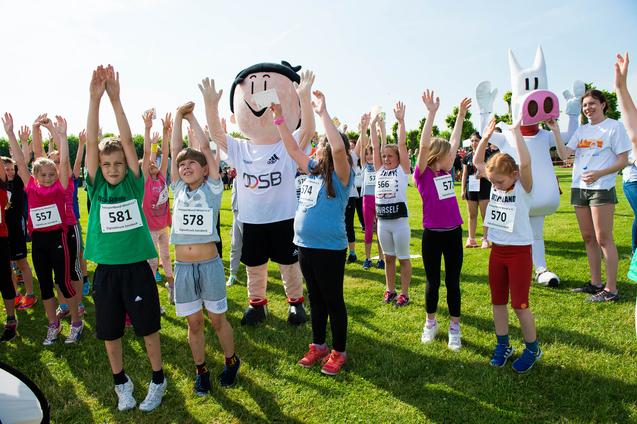 Kinder mit Nummern auf ihren T-Shirts stehen auf einer Wiese und heben die Arme, umgeben von Maskottchen.