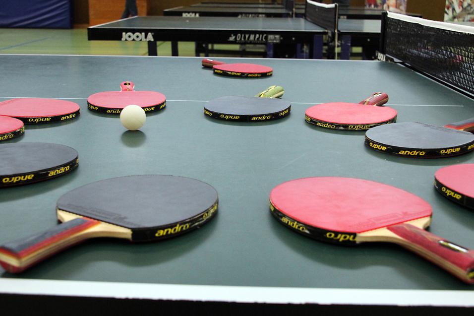 Mehrere Tischtennisschläger in verschiedenen Farben und ein Tischtennisball liegen auf einer grünen Tischtennisplatte.