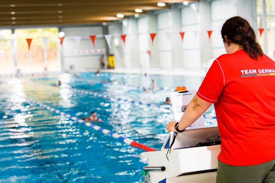 Schwimmbecken mit mehreren Schwimmern und einer Aufsichtsperson in einem roten Team-Germany-Shirt im Hintergrund.