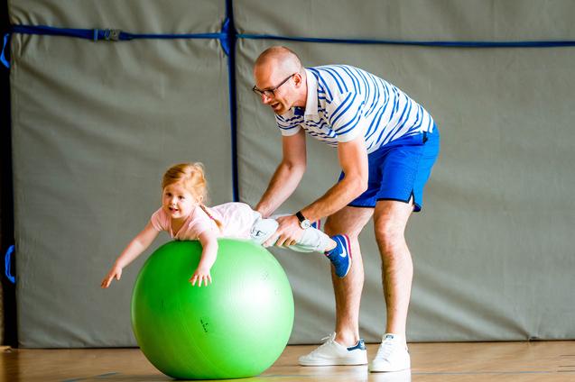 Ein Mann hält ein Kind, das auf einem grünen Gymnastikball sitzt, in einer Sporthalle. Beide lachen.