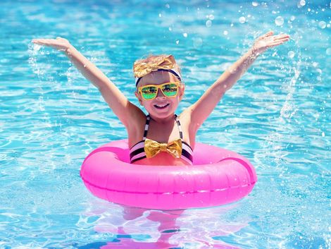 Ein Kind mit rosa Schwimmreifen schwimmt in einem Pool und hebt beide Arme nach oben.